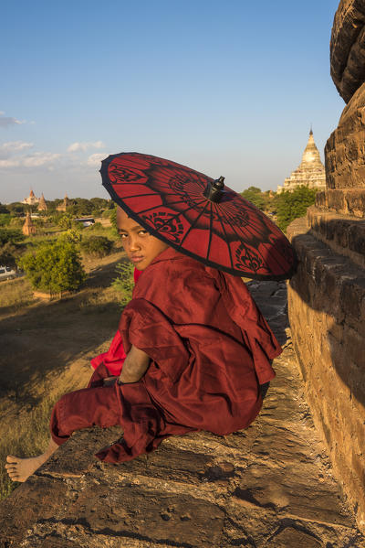 Bagan, Mandalay region, Myanmar (Burma). A young monk watching the camera with red umbrella sitting on top of a stupa.