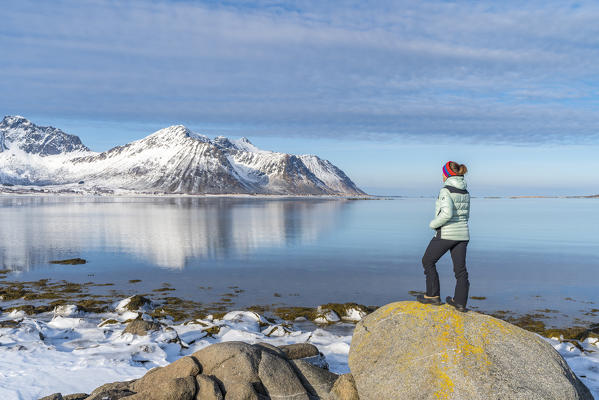 Woman standing on a rock admiring the Limstrandtinden snowcapped group in the background, in winter. Vestvagoy municipality, Lofoten district, Nordland county, Norway.