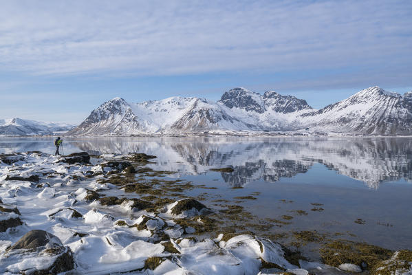 Man standing on a rock taking pictures of the Limstrandtinden snowcapped group in the background, in winter. Vestvagoy municipality, Lofoten district, Nordland county, Norway.
