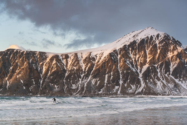 Man surfboarding in the sea in winter at sunset. Skagsanden beach, Flakstad, Nordland county, Northern Norway, Norway.