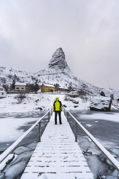 Man standing on a wooden bridge under the snow, looking towards Hammarskaftet peak. Reine, Nordland county, Northern Norway, Norway.