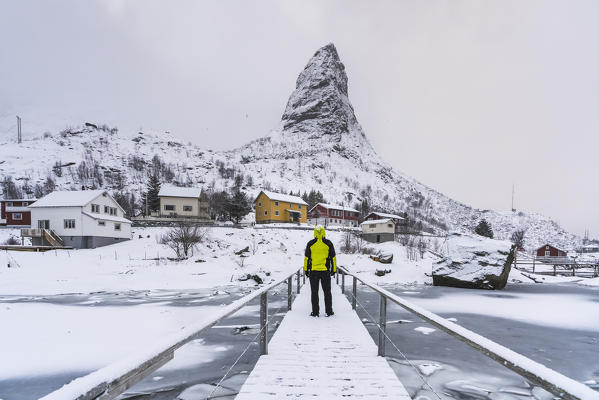 Man standing on a wooden bridge under the snow, looking towards Hammarskaftet peak. Reine, Nordland county, Northern Norway, Norway.