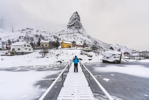 Woman standing on a wooden bridge under the snow, looking towards Hammarskaftet peak. Reine, Nordland county, Northern Norway, Norway.