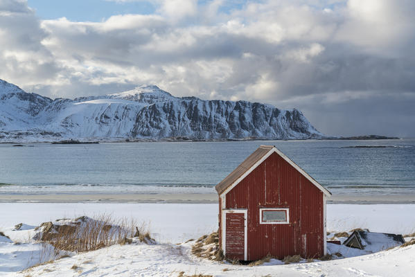 Red cabin in the snow on Ramberg beach, with mountains in the background. Flakstad municipality, Nordland county, Northern Norway, Norway.