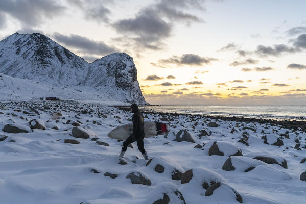 Surfer walking with his surfboard under the arm on Unstad Beach in winter at sunset. Vestvagoy municipality, Nordland county, Northern Norway, Norway.