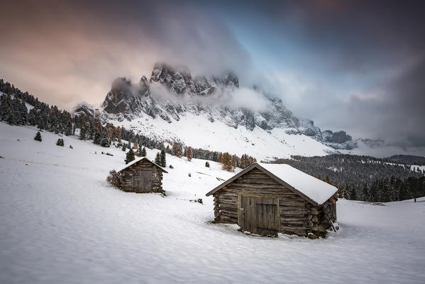 Winter scenery with two wooden lodges and Odle Dolomites covered in snow. Malga Gampen, Funes, Bolzano, Trentino Alto Adige - Sudtirol, Italy, Europe.