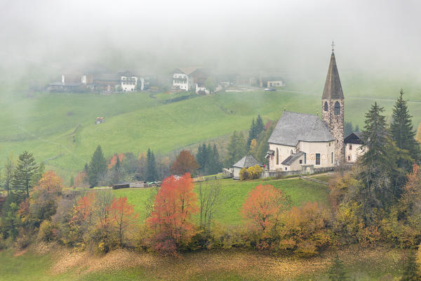 Church surrounded by autumnal trees and mist. Santa Maddalena, Funes, Bolzano, Trentino Alto Adige - Sudtirol, Italy, Europe.