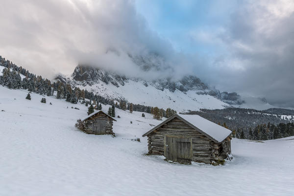 Winter scenery with two wooden lodges and Odle Dolomites covered in snow. Malga Gampen, Funes, Bolzano, Trentino Alto Adige - Sudtirol, Italy, Europe.