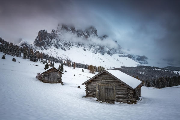 Winter scenery with two wooden lodges and Odle Dolomites covered in snow. Malga Gampen, Funes, Bolzano, Trentino Alto Adige - Sudtirol, Italy, Europe.