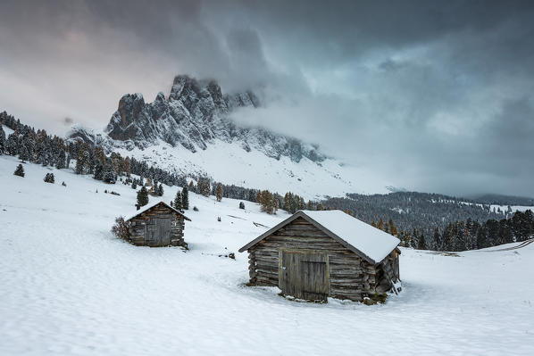 Winter scenery with two wooden lodges and Odle Dolomites covered in snow. Malga Gampen, Funes, Bolzano, Trentino Alto Adige - Sudtirol, Italy, Europe.