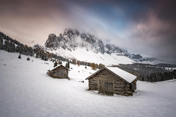 Winter scenery with two wooden lodges and Odle Dolomites covered in snow. Malga Gampen, Funes, Bolzano, Trentino Alto Adige - Sudtirol, Italy, Europe.