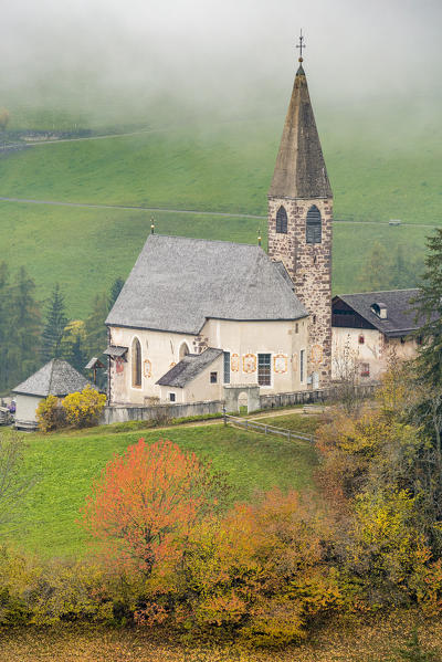 Church surrounded by autumnal trees and mist. Santa Maddalena, Funes, Bolzano, Trentino Alto Adige - Sudtirol, Italy, Europe.