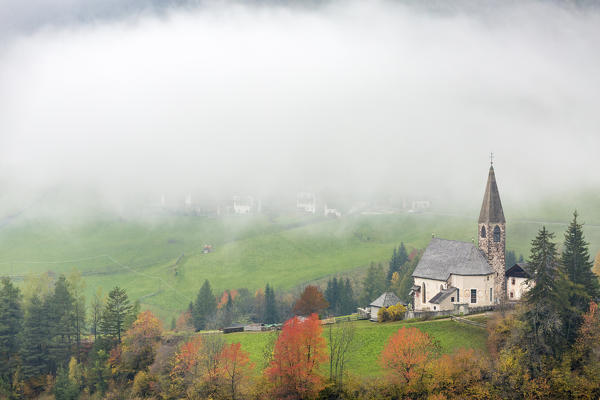 Church surrounded by autumnal trees and mist. Santa Maddalena, Funes, Bolzano, Trentino Alto Adige - Sudtirol, Italy, Europe.
