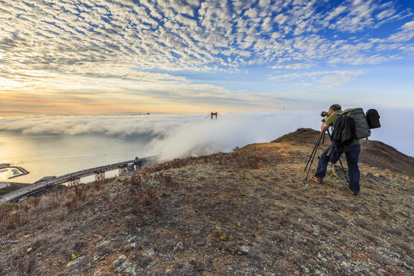 A photographer takes pictures of Golden Gate Bridge at sunrise from Slackers Hill. San Francisco, Marin County, California, USA.