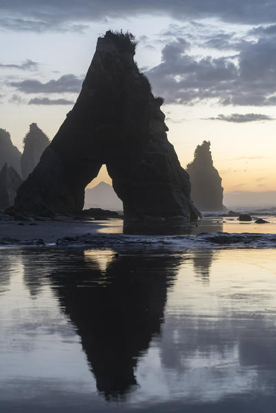 Rock reflect with low tide at the Three Sisters beach, at sunset, with Mt Taranaki seen through the rock cave.  Tongaporutu, New Plymouth district. Taranaki region, North Island, New Zealand.