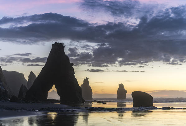 Rock reflect with low tide at the Three Sisters beach, at sunset, with Mt Taranaki seen through the rock cave. Tongaporutu, New Plymouth district. Taranaki region, North Island, New Zealand.