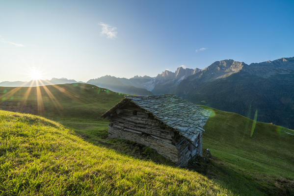 Stone hut in green pastures lit by warm sunburst at dawn, Tombal, Soglio, Val Bregaglia, canton of Graubunden, Switzerland