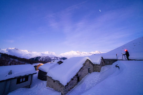 Hiker with head torch photographing the snowy huts of Groppera at dusk, Madesimo, Valchiavenna, Valtellina, Lombardy, Italy