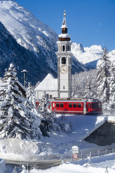 Winter sun over Bernina Express train and church bell tower of Bever village covered with snow, Graubunden canton, Switzerland
