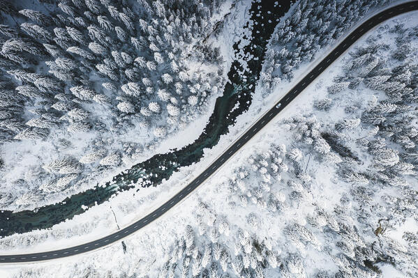 Car traveling on snowy mountain road across frozen river and woods, aerial view, Switzerland