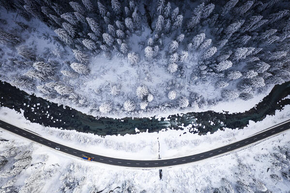 Snowplow on mountain road in the snowy woods on shore of frozen river, aerial view, Zernez, Engadin, Switzerland