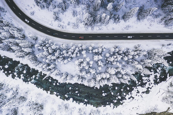 Aerial view of cars traveling in the forest covered with snow at the edge of frozen river in winter, Switzerland