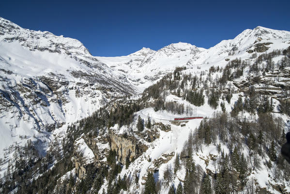 Aerial picture of the red Bernina train going to Bernina Pass in winter, Alp Grum, Poschiavo Valley, Canton Grigioni, Switzerland.