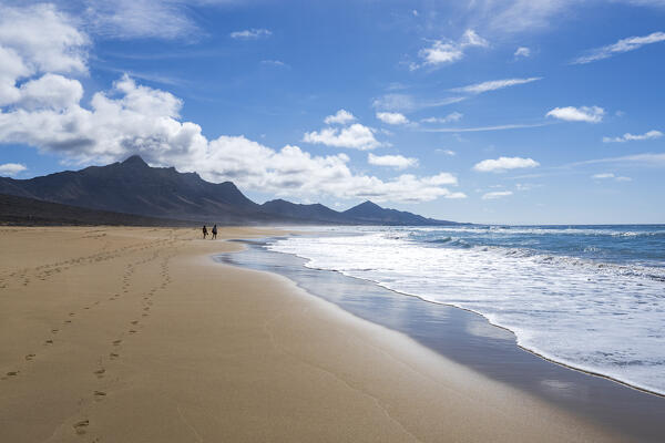 Man and woman walking on golden sand of Cofete beach washed by ocean waves, Jandia, Fuerteventura, Canary Islands, Spain