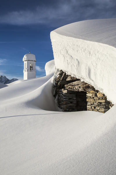 Mountain hut covered in snow with the bell tower of the Alpe Scima in the background. Valchiavenna, Valtellina Lombardy Italy Europe