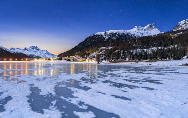 Starry sky over the snowy mountains and Silvaplana village seen from the frozen lake Champfer, Graubunden, Engadin, Switzerland