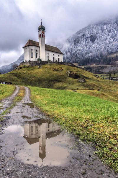 The Church of Wassen is reflected in a puddle after a winter snowfall. Andermatt. Canton of Uri. Switzerland. Europe
