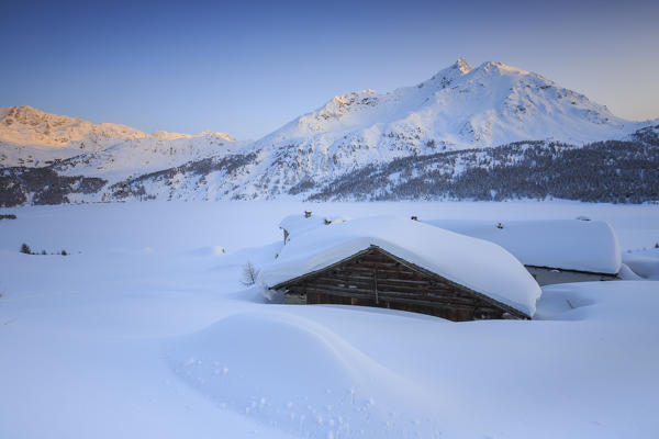 Spluga huts surrounded by snow with the sun illuminating the snowy peaks before disappearing into the horizon. Maloja Pass. Engadine. Switzerland. Europe