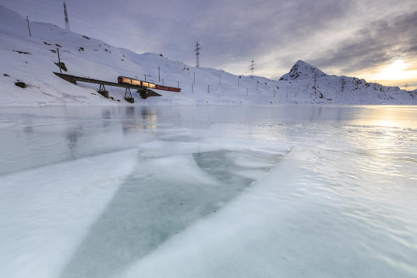 The Red Train passes on the shores of Lake Bianco at Bernina Pass completely frozen. Canton of  Graubunden. Engadine. Switzerland. Europe