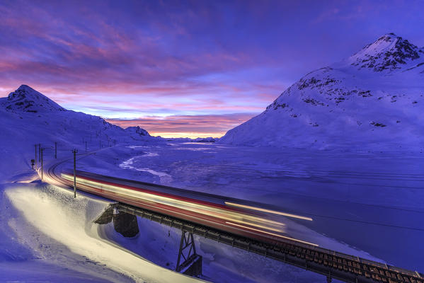 The Bernina Express passes with lights on close to the banks of Lake Bianco completely frozen with the sky illuminated by the colors of dusk. Canton of  Graubunden. Engadine. Switzerland. Europe