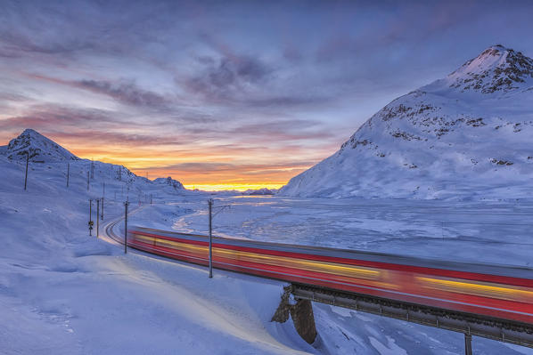 The Bernina Express passes on the shores of Lake Bianco completely frozen before the sun rises. Canton of  Graubunden. Engadine. Switzerland. Europe