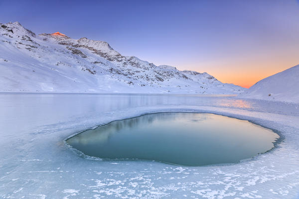 Soft colors of a cold dawn reflected in a pool of turquoise water surrounded by the snow in the middle of White Lake. Bernina Pass. Canton of Graubuenden. Engadine. Switzerland. Europe