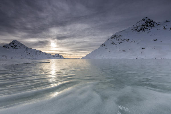 The sun's rays illuminate the frozen surface of White Lake where the snow is hard to settle because of the constant wind that blows from the north. Bernina Pass. Canton of Graubuenden. Engadine. Switzerland. Europe