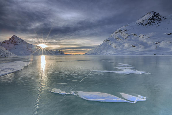 The sun's rays illuminate the frozen surface of White Lake where the snow is hard to settle because of the constant wind that blows from the north. Bernina Pass. Canton of Graubuenden. Engadine. Switzerland. Europe