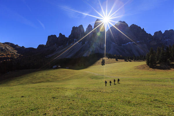 Hikers crossing the green pastures of Malga Glatsch illuminated by sunbeams. Funes Valley South Tyrol Dolomites Italy Europe