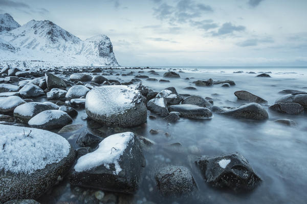 Rocks in the cold sea and snow capped mountains under the blue light of dusk Unstad Lofoten Islands Northern Norway Europe