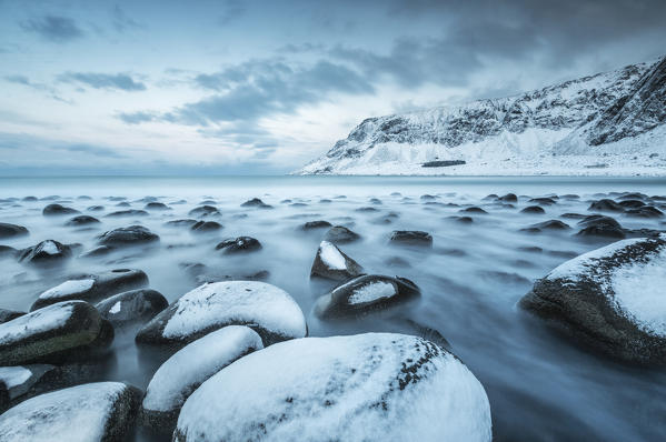 Rocks in the cold sea and snow capped mountains under the blue light of dusk Unstad Lofoten Islands Northern Norway Europe