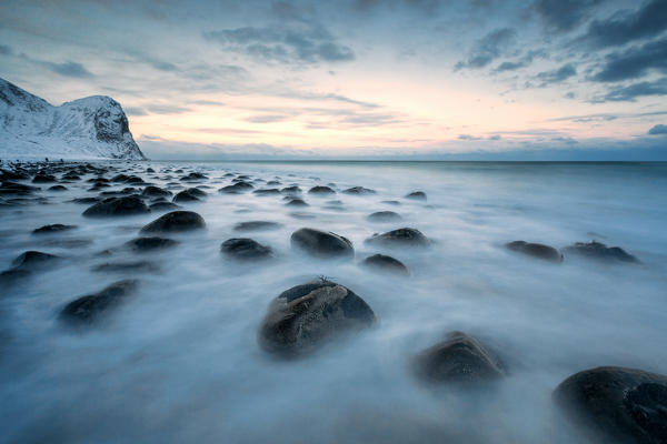 Rocks in the cold sea and snow capped mountains under the blue light of dusk Unstad Lofoten Islands Northern Norway Europe