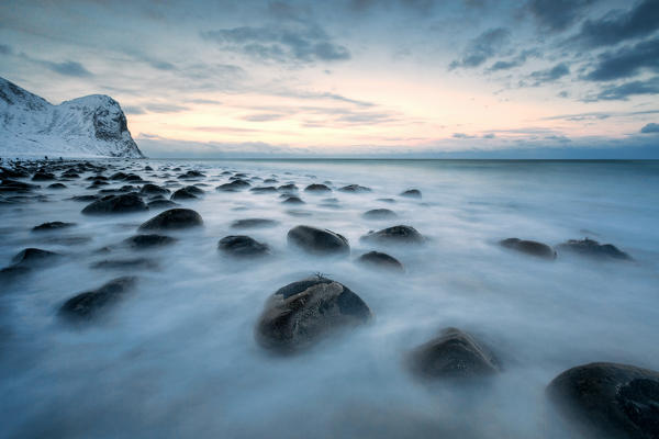 Rocks in the cold sea and snow capped mountains under the blue light of dusk Unstad Lofoten Islands Northern Norway Europe