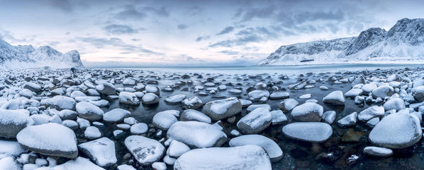 Rocks in the cold sea and snow capped mountains under the blue light of dusk Unstad Lofoten Islands Northern Norway Europe