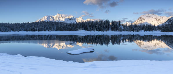 Wooden cabin surrounded by snowy peaks and woods reflected in Palù Lake at dawn Malenco Valley Valtellina Lombardy Italy Europe