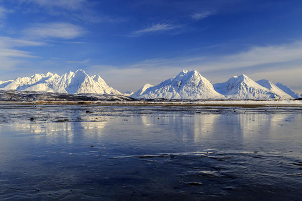 The blue sky and snow capped mountains are reflected in the frozen water 
Breivikeidet Lyngen Alps Tromsø Lapland Norway Europe