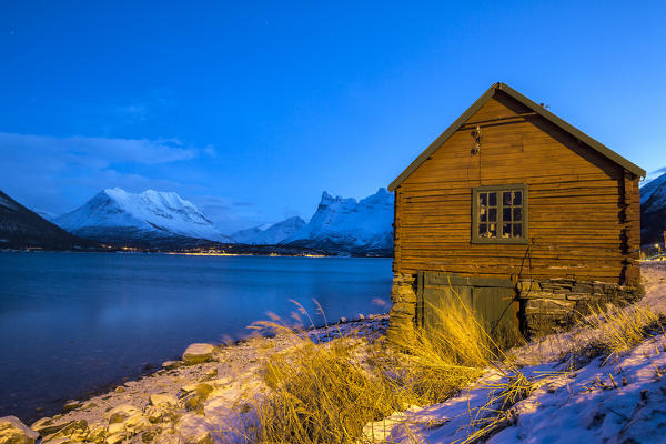 Wooden cabin of fisherman illuminated surrounded by the frozen sea at dusk Storfjorden Lapland Lyngen Alps Tromsø Norway Europe