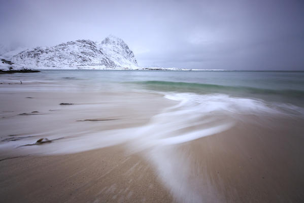 Waves of the icy sea on the beach in the background the snowy peaks Pollen Vareid Flakstad Lofoten Islands Norway Europe