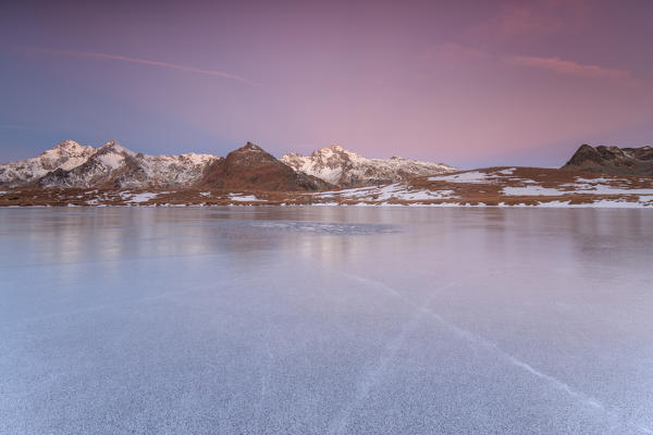 The snowy peaks are reflected on the frozen surface of Andossi Lake at dawn Spluga Valley Valtellina Lombardy Italy Europe