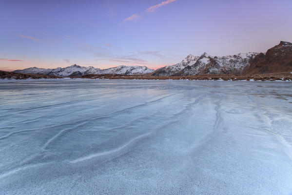 The lights of dawn on the snowy peaks around the frozen surface of Andossi Lake Spluga Valley Valtellina Lombardy Italy Europe